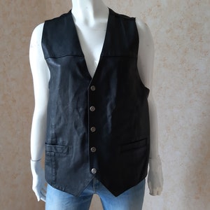 May include: A black leather vest with a V-neck and a button-up front. The vest has two front pockets and is displayed on a mannequin. The vest is worn over a light blue denim.