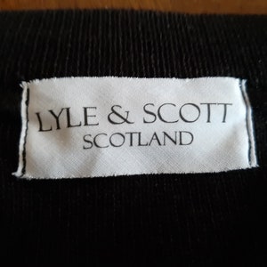 May include: Close-up of a black fabric with a white label. The label reads "LYLE & SCOTT SCOTLAND" in black capital letters. The fabric has a ribbed texture.