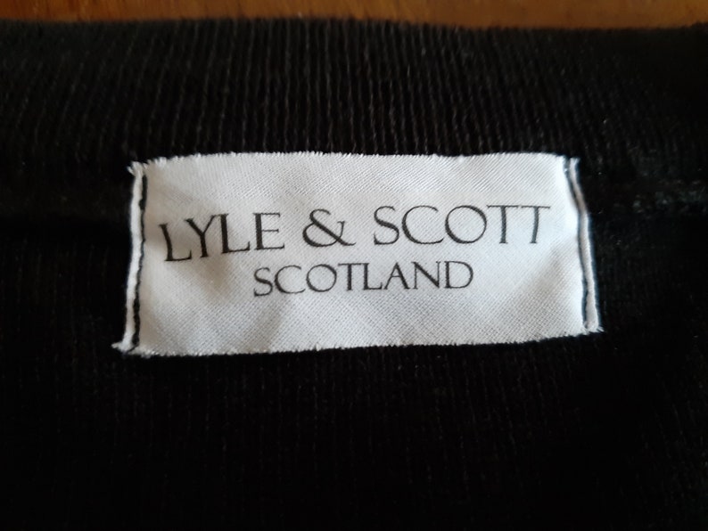 May include: Close-up of a black fabric with a white label. The label reads "LYLE & SCOTT SCOTLAND" in black capital letters. The fabric has a ribbed texture.
