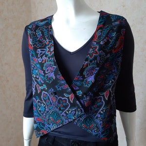 Puede incluir: Un chaleco sin mangas con base negra y un vibrante estampado de cachemira multicolor en tonos azul, morado y rojo. El chaleco presenta un escote en V, un cierre frontal cruzado con pequeños botones negros y un dobladillo corto y angular.