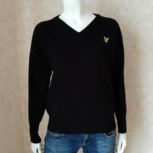 May include: A black V-neck sweater with long sleeves, displayed on a mannequin. A small yellow embroidered bird logo is on the left chest. The sweater is paired with blue jeans.