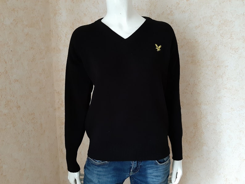 May include: A black V-neck sweater with long sleeves, displayed on a mannequin. A small yellow embroidered bird logo is on the left chest. The sweater is paired with blue jeans.