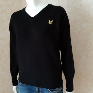 May include: Black V-neck sweater with long sleeves. A small yellow bird emblem is embroidered on the left chest. The sweater is displayed on a mannequin. The background is a light beige color.