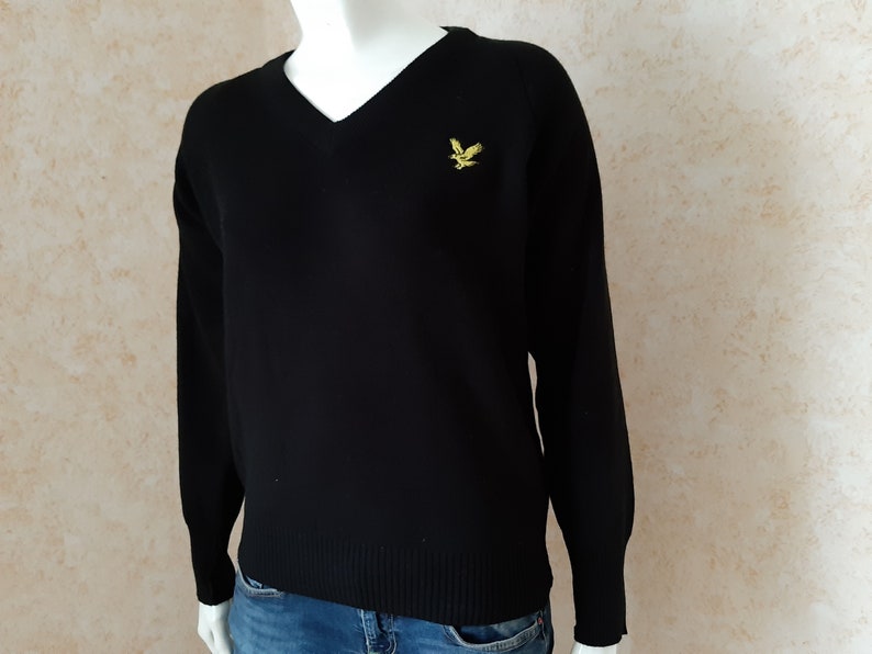 May include: Black V-neck sweater with long sleeves. A small yellow bird emblem is embroidered on the left chest. The sweater is displayed on a mannequin. The background is a light beige color.