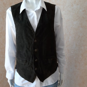 Può includere: Un gilet in camoscio marrone scuro con scollo a V, quattro bottoni metallici rotondi sul davanti e due piccole tasche a filetto in vita. Il gilet è indossato sopra una camicia bianca a maniche lunghe con colletto e chiusura a bottoni.