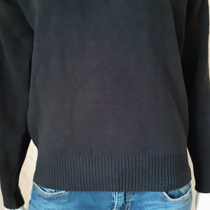 May include: A black sweater with long sleeves and a ribbed hem. The sweater is made of a soft material and is worn with blue jeans. The overall style is casual and comfortable.
