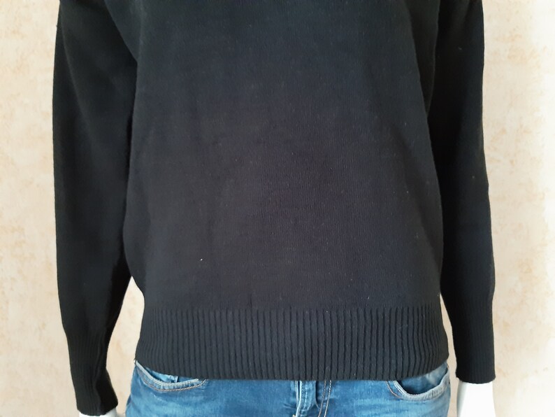 May include: A black sweater with long sleeves and a ribbed hem. The sweater is made of a soft material and is worn with blue jeans. The overall style is casual and comfortable.