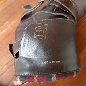 Pu&ograve; includere: Stivale in pelle marrone con cinturini con fibbia e un dettaglio in tessuto a quadri alla base. Lo stivale ha una finitura lucida e il testo "MADE IN TUNISIA" &egrave; visibile. Lo stivale &egrave; appoggiato su una superficie di legno.