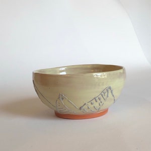 May include: A ceramic bowl with a glossy cream-coloured glaze and a rounded shape. The exterior features a hand-drawn mountain range design in light blue, while the base of the bowl is exposed, unglazed terracotta-coloured clay.