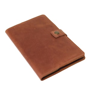 May include: A brown leather notebook with a flap closure and a brass button. The notebook is closed and lying flat on a white surface.