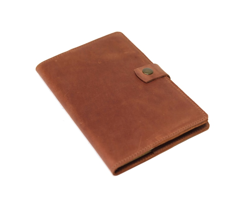 May include: A brown leather notebook with a flap closure and a brass button. The notebook is closed and lying flat on a white surface.