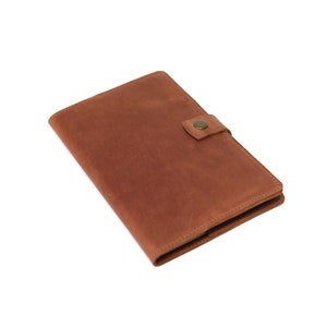 May include: A brown leather notebook with a flap closure and a button. The notebook is closed and lying on a white surface.