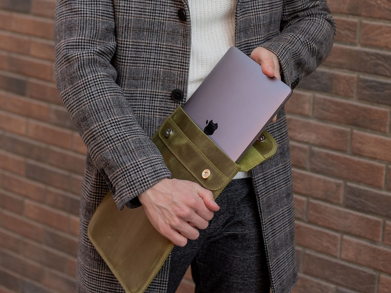 May include: A person holding a green leather laptop sleeve with a silver laptop inside. The sleeve has a flap with a button closure.