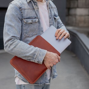 May include: A person holding a brown leather laptop sleeve with a silver laptop inside. The sleeve has a flap that closes with a magnetic snap.
