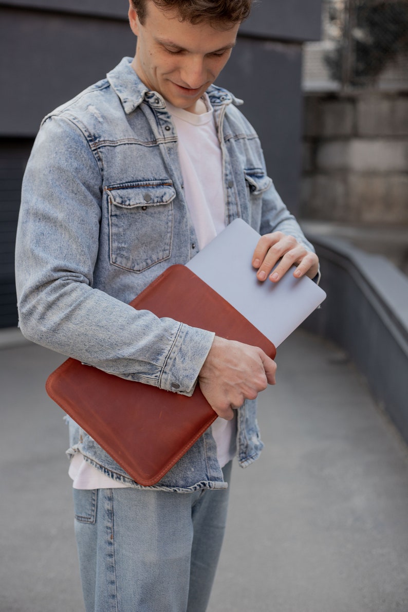 May include: A person holding a brown leather laptop sleeve with a silver laptop inside. The sleeve has a flap that closes with a magnetic snap.