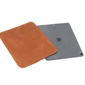 May include: A brown leather sleeve for a tablet computer. The sleeve is folded over the tablet, which is gray and has a silver Apple logo.