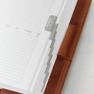 May include: A brown leather bound planner with a white page open to May 6th and 7th. The planner has a tab for each day of the month. The tab for May 7th is visible and says "ТРАВЕНЬ" which is Ukrainian for "May".