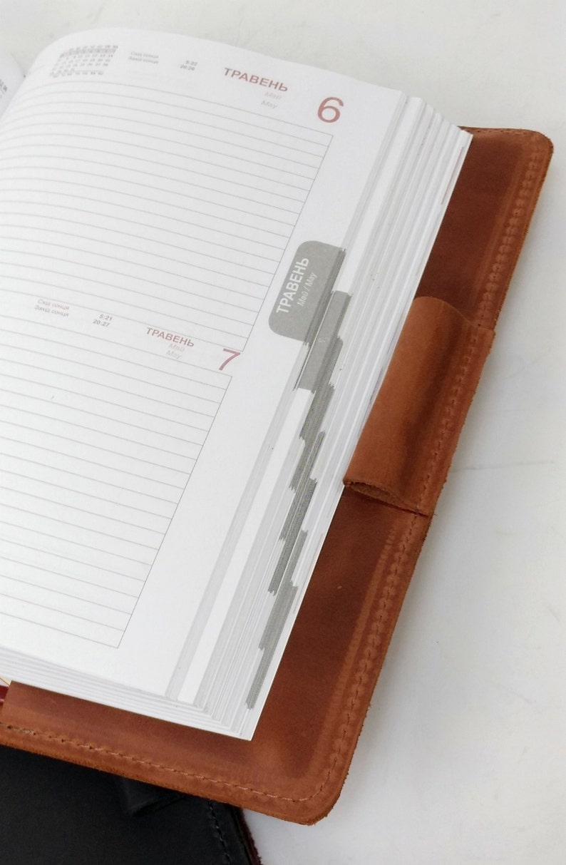 May include: A brown leather bound planner with a white page open to May 6th and 7th. The planner has a tab for each day of the month. The tab for May 7th is visible and says "ТРАВЕНЬ" which is Ukrainian for "May".