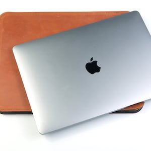 May include: A silver laptop computer with an Apple logo on the lid is closed and sitting inside a brown leather sleeve.