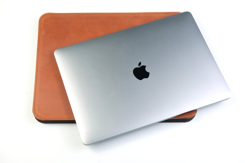 May include: A silver laptop computer with an Apple logo on the lid is closed and sitting inside a brown leather sleeve.