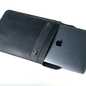 May include: A black leather laptop sleeve with a flap closure and two snap buttons. The sleeve is open and a silver laptop is partially inside.