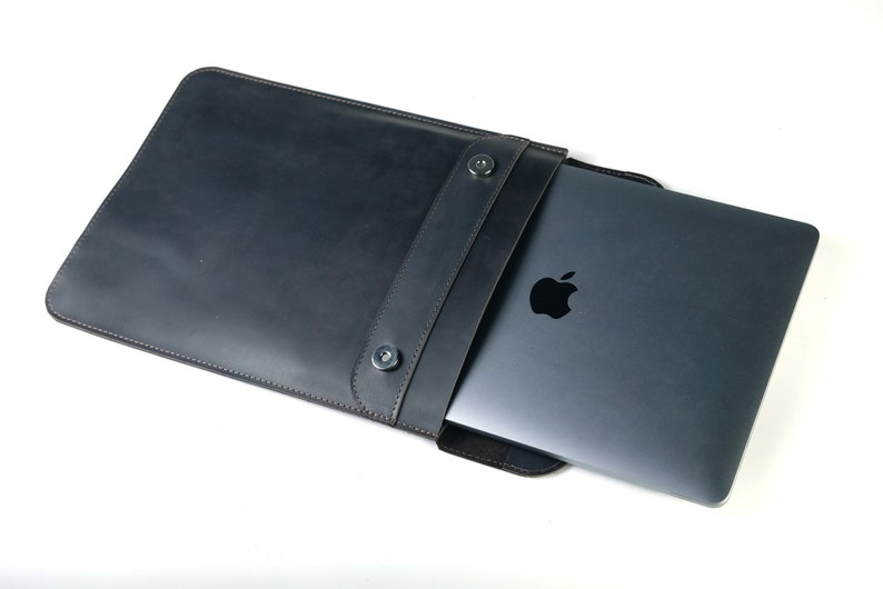 May include: A black leather laptop sleeve with a flap closure and two snap buttons. The sleeve is open and a silver laptop is partially inside.