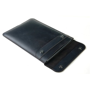 May include: A dark blue leather laptop sleeve with a flap closure and snap buttons. The sleeve is open to reveal a soft black interior.