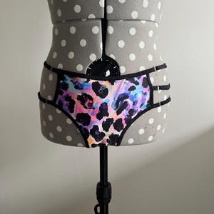 May include: Low-rise underwear featuring a vibrant, multicolored leopard print pattern in shades of pink, blue, and orange. The garment is trimmed with black fabric and includes thin, adjustable black side straps with silver-toned metal hardware.