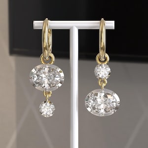 Puede incluir: Un par de pendientes de aro dorados con un diamante grande de talla ovalada y un diamante redondo más pequeño colgando. Los pendientes se exhiben en un soporte blanco, mostrando su brillo.
