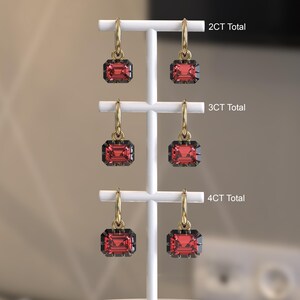 May include: Three pairs of gold hoop earrings with emerald-cut red gemstones hanging from a white display stand. The pairs are labeled 2CT Total, 3CT Total, and 4CT Total, indicating the total carat weight of the red stones in each set of earrings.
