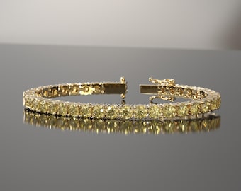 May include: A gold tennis bracelet with a row of yellow gemstones. The bracelet is open, showing the clasp mechanism. The gemstones are evenly spaced and reflect light, creating a sparkling effect. The bracelet is displayed on a reflective surface.