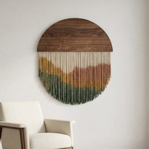 May include: A circular wall hanging featuring a dark brown wooden semi-circle top piece with a natural wood grain. Attached to the bottom is a dense fringe of vertical cotton cords dyed in layered horizontal bands of cream, mustard, and dark green.