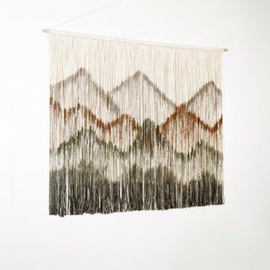 May include: A woven wall hanging with a mountain design. The artwork features a cream-colored background with brown, green, and gray mountain shapes. The piece is suspended from a wooden dowel and has long, fringed threads.