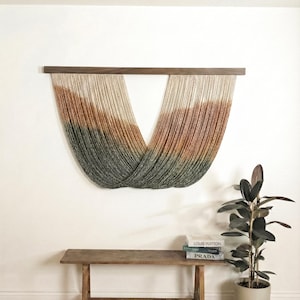 May include: A decorative wall hanging with a wooden top bar, composed of cascading strands in beige, brown, and green. The design is curved and flowing. A wooden bench and a potted plant are visible in the foreground.
