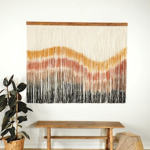 May include: A large wall hanging made of yarn with a wavy pattern in shades of black, brown, orange, and yellow. The hanging is attached to a wooden dowel rod.