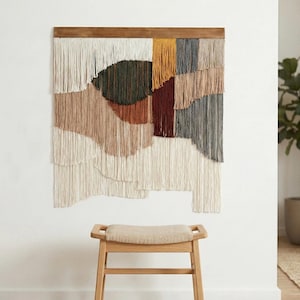 May include: A large, rectangular wall hanging with a wooden frame. The artwork features layered, fringed textiles in shades of cream, brown, green, and gray. The piece is mounted above a light-colored wooden stool with a beige cushion.