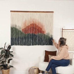 May include: A large wall hanging with a sunset scene in shades of orange, red, green, and blue. The hanging is made of yarn and is attached to a wooden dowel.