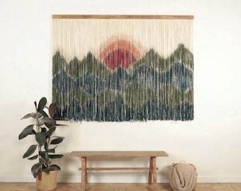 May include: A large rectangular wall hanging made of cream-coloured fibre strands suspended from a wooden rod. The fibres are dyed to create a landscape scene featuring a red sun rising over layered mountain ranges in shades of green and blue.