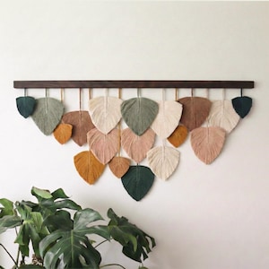 May include: A macrame wall hanging featuring leaf-shaped designs in various colors, including green, pink, and beige. The leaves are suspended from a dark brown wooden rod, creating a decorative and textured wall art piece.