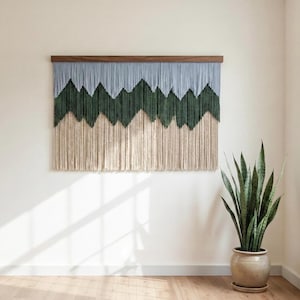 Può includere: Un grande arazzo da parete con un motivo a montagna. La sezione superiore è di colore azzurro, con una catena montuosa verde scuro al centro e una frangia beige nella parte inferiore. L'arazzo da parete è montato su una barra di legno.