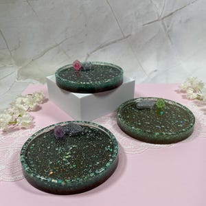 May include: Three round green glitter resin coasters with a clear epoxy top. Each coaster has a small pink, green, or purple gem embedded in the resin.