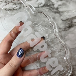 May include: Clear acrylic bookmark with the words "Chapter" and "Just one more" etched in a cursive font. The bookmark has a cloud-like shape with a small hole at the top. The background is a marble pattern.