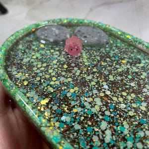 May include: A green and blue glitter resin tray with two grey stones and a pink pig figurine.