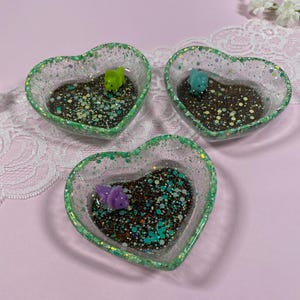 May include: Three heart-shaped resin trinket dishes with green glitter edges. Each dish has a brown and white speckled base with green, blue, and white glitter accents. A small green, blue, and purple figurine is in each dish.
