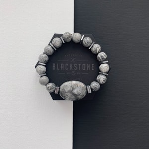 May include: A gray and white stone bracelet with silver accents. The bracelet is on a black and white background. The bracelet has the text "AUTHENTIC BLACKSTONE EST. 2013 HAND CRAFTED" on it.