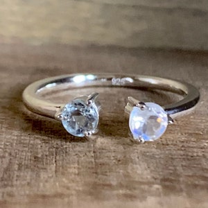 May include: A silver ring with two stones. The stone on the left is a light blue aquamarine, and the stone on the right is a white moonstone.