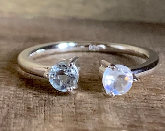 May include: A silver ring with two stones. The stone on the left is a light blue aquamarine, and the stone on the right is a white moonstone.