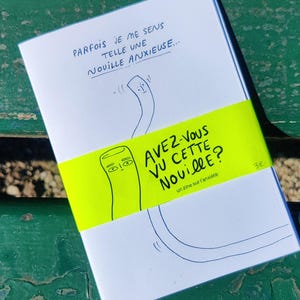 Peut inclure: Un livret blanc avec des dessins au trait bleus et noirs et du texte. La couverture porte l'inscription "PARFOIS JE ME SENS TELLE UNE NOUILLE ANXIEUSE..." et un dessin de nouille. Une bande jaune fluo entoure le livret avec le texte "AVEZ-VOUS VU CETTE NOUILLE?" et "un zine sur l'anxiété".