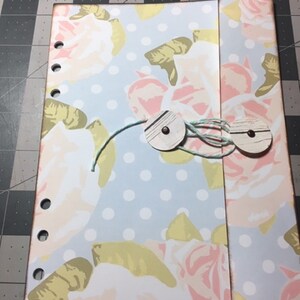 May include: A light blue and white floral patterned notebook with a pocket on the right side. The notebook has a polka dot pattern and is decorated with two wooden buttons and a light blue string.