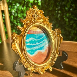 Puede incluir: Una pequeña pintura ovalada de una escena de playa con olas azules y una playa de arena, enmarcada en un marco dorado adornado con un soporte negro.